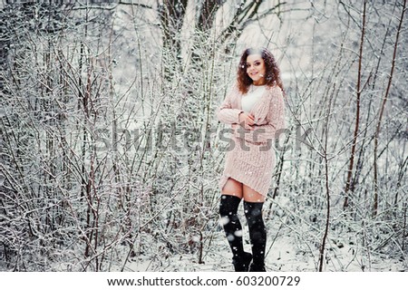 Curly brunette girl background falling snow, wear on warm knitted sweater, black mini skirt and wool stockings. Model on winter. Fashion portrait at snowy weather. toned photo.