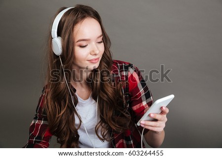 Picture of beautiful young lady posing isolated over grey background listening music with headphones chatting by phone. Looking aside.