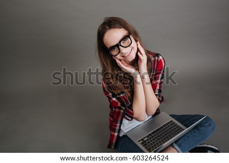 Image of incredible smiling young lady wearing glasses sitting isolated over grey background chatting by laptop computer. Looking at camera.