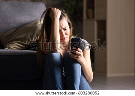 Front view of a sad teen checking phone sitting on the floor in the living room at home with a dark background Royalty-Free Stock Photo #603280052