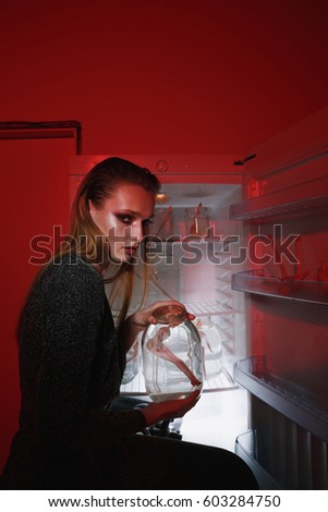 Side view of woman in dress which sitting near the fridge and holding jar with doll while looking at camera. Vertical image. Conceptual picture