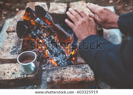 Traveler warming his hands by the campfire outdoors. Tea or coffee in aluminum mug on background. Camping detail, travel lifestyle photo.