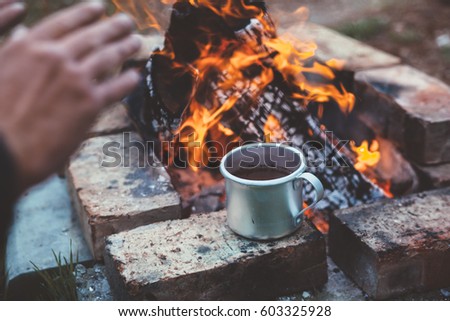Traveler warming his hands by the campfire outdoors. Tea or coffee in aluminum mug on background. Camping detail, travel lifestyle photo.