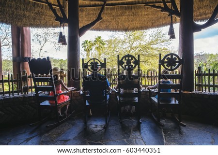 Rear view of young family sitting together on the thatched roof balcony of their tropical island hotel. People sitting in rocking chairs are silhouetted against the scenic island setting.