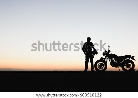 Biker man and motorcycle,Young man rider trendy motorbik had stopped to rest during the trip to see the light of nature,light of the sky between change day to night. silhouette wallpaper concept Royalty-Free Stock Photo #603510122