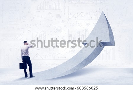 Young sales business person in elegant suit standing with his back in front of a big arrow pointing up and a clear background full of pie charts, numbers