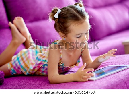 Little preschool girl lying on couch in living room using tablet play online games download interesting applications watching cartoons videos in internet spending free time alone at home 