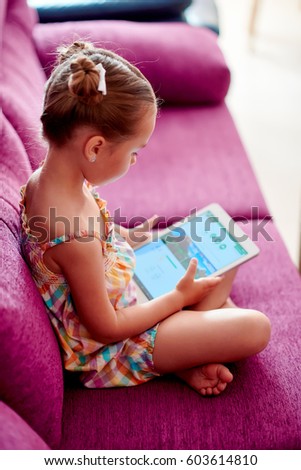 Preschool cute girl little daughter using tablet computer sitting on a sofa at home downloading choosing new game, play online,  internet user, generation addicted with modern gadgets devices concept