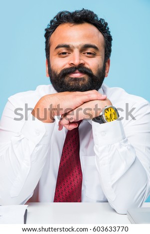 portrait of young Indian businessman in beard, sitting in relaxed position at at office table with pleasant look. Confident asian male business person