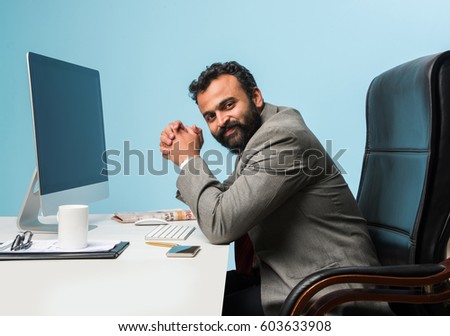 Indian/asian businessman in beard, sitting in relaxed position at at office table with computer having pleasant look