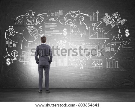 Rear view of a businessman in a gray suit looking at business scheme drawn on a blackboard. Concept of a business plan.