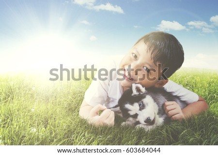 Picture of cheerful boy embracing husky puppy while lying in the meadow and looking at the camera