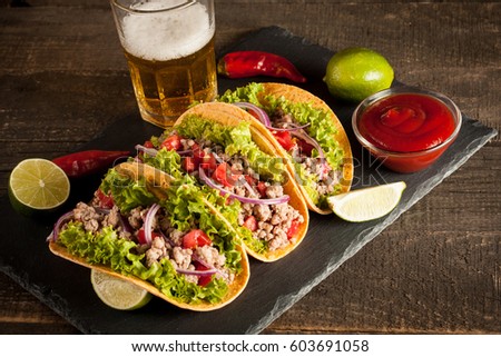 Photo of Mexican tacos with ground meat, beef, beans, onions and salsa on wooden background. Ketchup sauce and lime. A glass o beer in the background.