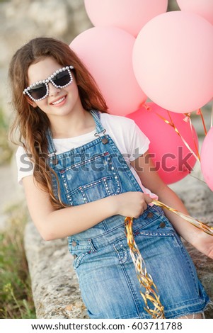 Happy little girl with balloons summer portrait. Hipster look child girl 10 year old with sunglasses and balloons wear jeans dress and posing outdoors at sunset. Cute kid friendly smiling outdoors.