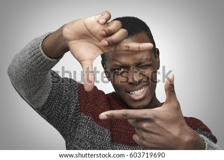 Close up portrait of young African American male in warm casual sweater making frame with his hands, looking at camera and smiling, standing against isolated grey background. Dark-skinned photographer