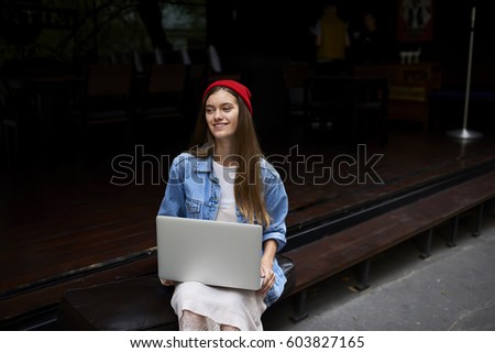 Charming stylishly dressed young female IT developer enjoying doing remote job sitting outdoors in cafe with laptop computer using free wireless connection to internet while sharing multimedia files