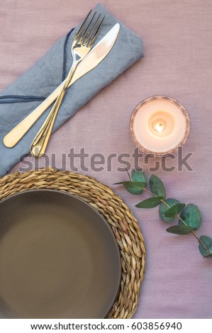 Elegant table setting on purple linen cloth dark plate rattan coaster cutlery blue napkin, burning candle, twig of silver dollar eucalyptus, top view, flat lay, styled stock photo