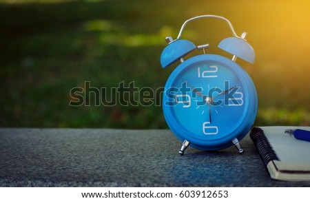 Blue alarm clock and notebook on a wooden desk with blurred nature background.