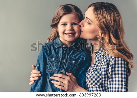 Cute little girl and her beautiful young mom on gray background. Mom is kissing her daughter in cheek