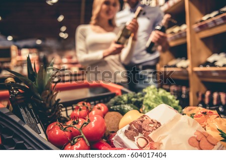 At the supermarket. Shopping cart full of goods, in the background young couple choosing wine Royalty-Free Stock Photo #604017404