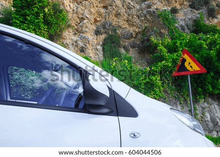 Contextual image concept: insurance - auto, property and life. Road sign falling stones, Traffic sign caution possible falling rocks from the mountains. Car standing under rock in a dangerous place.