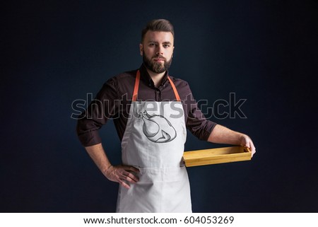 Young bearded man, waiter, chef, dressed in white apron with picture of tomatoes,standing,holding in one hand blank wooden tray and looking at camera.Image of roasted turkey.