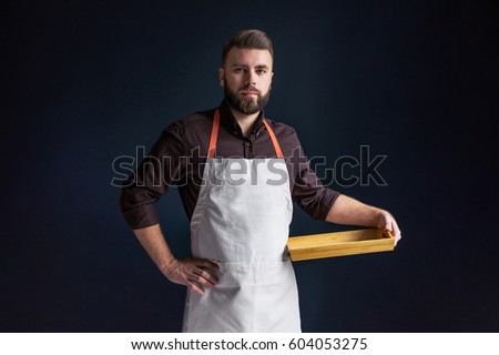 Young bearded man, waiter, chef, dressed in white apron with picture of tomatoes,standing,holding in one hand blank wooden tray and looking at camera.