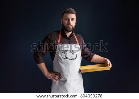 Young bearded man, waiter, chef, dressed in white apron with picture of tomatoes,standing,holding in one hand blank wooden tray and looking at camera.The image of vegetables.