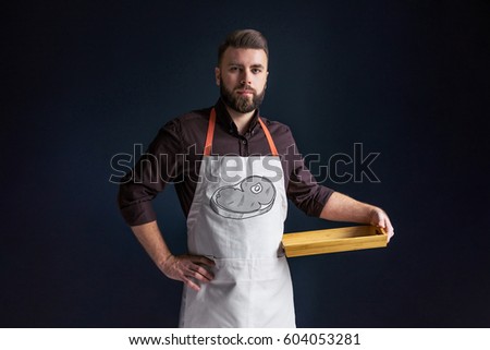 Young bearded man, waiter, chef, dressed in white apron with picture of tomatoes,standing,holding in one hand blank wooden tray and looking at camera.The picture of the steak.