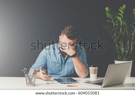 Front view. Young bearded sad businessman is sitting at table, covering his face with his hand and holding smartphone in his hand.On desk is laptop, tablet computer. Man got bad news.Instagram filter.