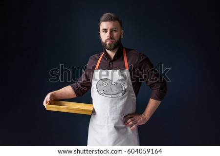 Front view. Young bearded man, waiter, chef, dressed in white apron with picture of meat steak,standing,holding in one hand blank wooden tray and looking at camera. The picture of the steak.
