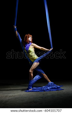 Beautiful young athletic girl, with redhead in yellow costume, posing with gorgeous blue aerial silks on the light in the darkness. Diagonal with canvases wrapped around slender legs.