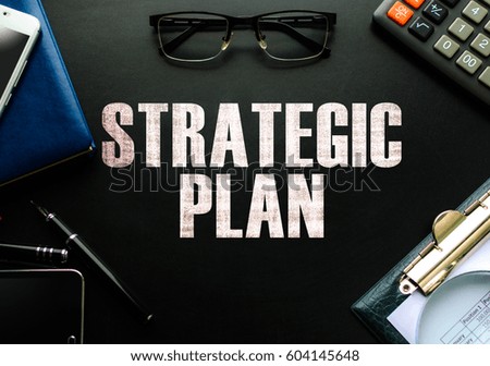 Black chalkboard with business accessories (notepad, magnifying glass, fountain pen, tablet, phone, glasses and calculator) and text STRATEGIC PLAN. Top view.