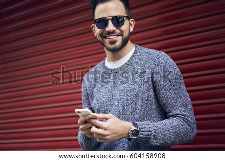 Handsome young male tourist in casual trendy sweatshirt typing messages in social networks via smartphone connected to 5G wireless standing on red background copy space area for your advertising