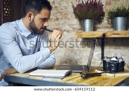 Professional creative bearded male journalist thinking over new report editing photo images made by vintage camera making illustration in retro style sitting in modern interior cafe using laptop 