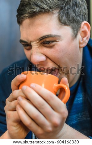 portrait of a young man - a teenager, full face , scar on eyebrow , pimples on the face , dressed in a blue hoodie and blue background . drinking tea from orange Cup