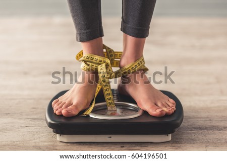 Cropped image of woman feet standing on weigh scales, on gray background. Legs winded with a tape measure Royalty-Free Stock Photo #604196501