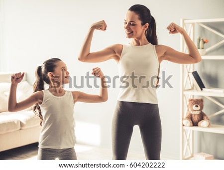 Beautiful young woman and charming little daughter are showing their biceps and smiling while working out at home Royalty-Free Stock Photo #604202222
