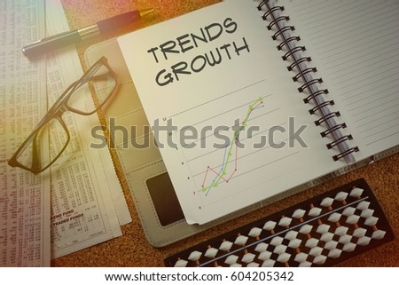 Notebook with writing (TRENDS GROWTH) and upward colored graphs, newspapers, pen, glasses and abacus on a wooden table, view from above. Financial concept