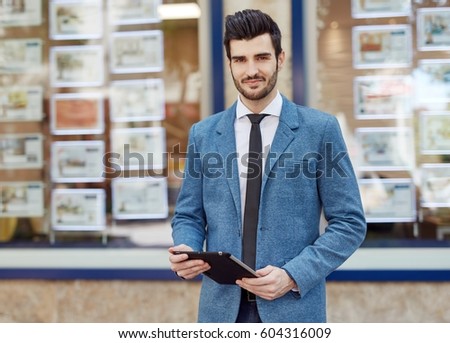 Portrait of handsome young real estate agent standing front of agency, holding tablet.