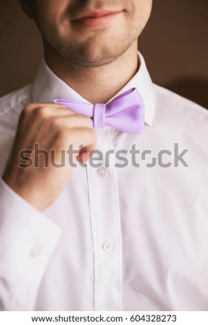 Elegant groom in white shirt holds stylish lilac bow tie in his hands.