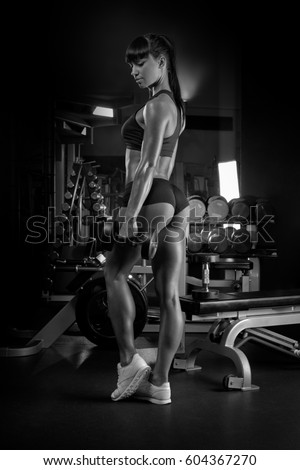 Black and white image of muscular young woman athlete standing looking down with her hands with dumbell. Woman bodybuilder relaxing after exercise in gym.