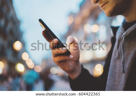 Side view image of young hipster man using modern smartphone outdoors, man typing an sms message while walking at evening city streets, social networks concept