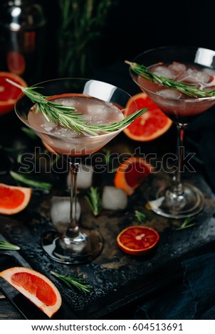 Two glasses with a three-layered grapefruit and rosemary alcoholic cocktail, slices of grapefruit and ice cubes on a black background. Toned image. Close up and selective focus