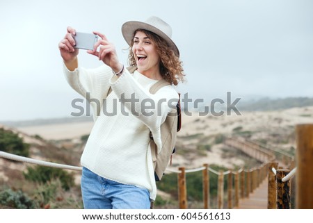 Happy woman with hat and backpack taking photo with her smartphone, traveling among natural sand meadows