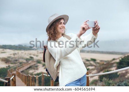 Handsome woman with hat and backpack taking photo with her smartphone, traveling among beautiful sand meadows