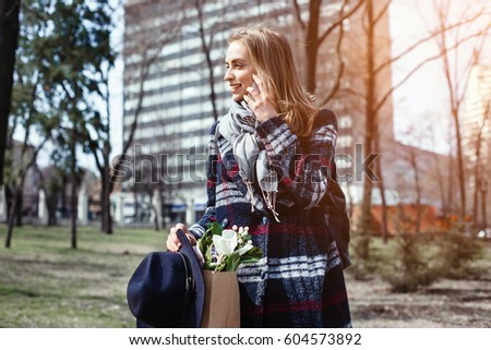 Young woman talking with parents on cell telephone walking on a sunny day in the park. Attractive girl in a coat, hat and flowers in the park in the spring