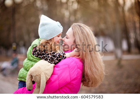 Blonde mom and little daughter in pink hats and jackets smile, hug and laugh outdoors in the spring for a walk in the park