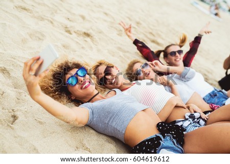 Side view. Happy girlfriends taking selfies lying on sand. Smiling happily. Cool trendy summer outfit.