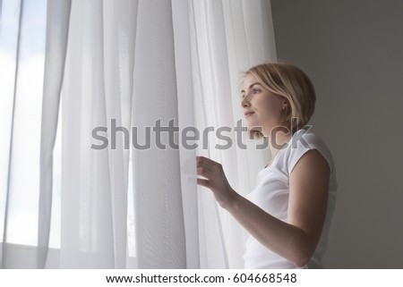 Young pretty smiling girl opening white curtains in morning to enjoy blue sky